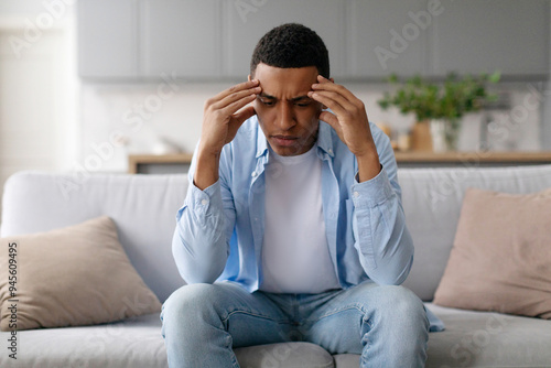 Stressed young black man sitting on couch at home, rubbing temples, suffering from headache migraine or thinking