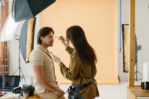 Stylist woman applying make up to handsome model at photo shoot backstage, getting ready for professional photoshoot.