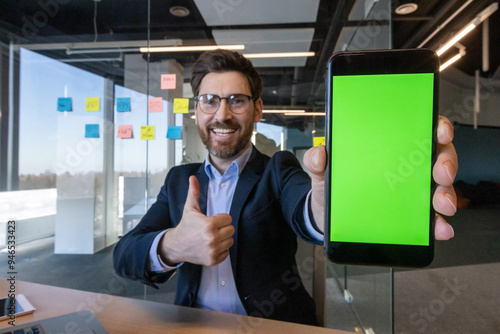 Businessman in modern office shows smartphone with green screen, expressing positivity with thumbs up gesture. Ideal for technology, success, and professional concepts in corporate environment.