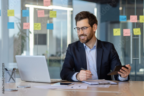 Businessman in a suit working in modern office. Smiling while using a calculator and laptop, surrounded by sticky notes on glass wall. Represents accounting, finance, and business planning.