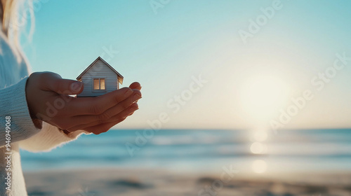 Female hands cradling a small house, with the ocean and sandy beach in the background, representing a serene coastal retreat.