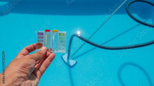 Hand holding a pool water testing kit above a clear blue pool with cleaning equipment in the background.