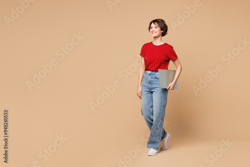 Full body side view young happy smart fun IT woman wear red t-shirt casual clothes hold closed laptop pc computer walk go isolated on plain pastel light beige background studio . Lifestyle concept.