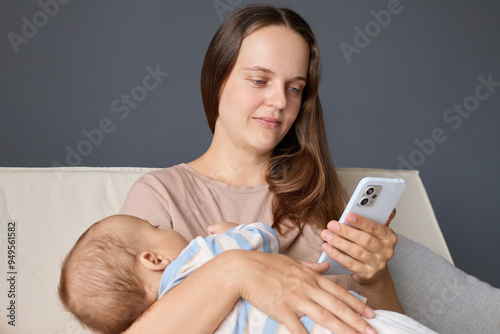 Brown haired mother at home with her infant browsing the internet on her smartphone managing communications through various apps and taking care of infant daughter or son