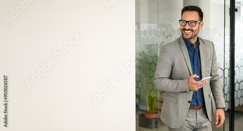 Confident male business leader texting over smart phone and looking away while standing by white wall