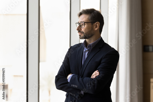 Thoughtful serious company executive man in formal jacket and glasses standing at office window with hands folded, looking away, thinking on real estate investment, future project