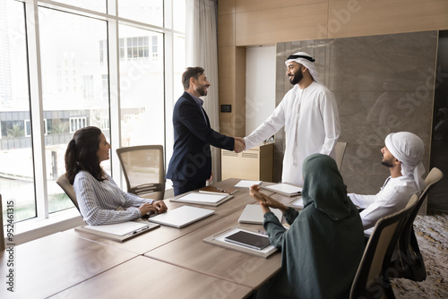 Positive confident Arab and European businessmen shaking hands at team meeting, standing at large table, talking, smiling, laughing, discussing contract, deal, partnership