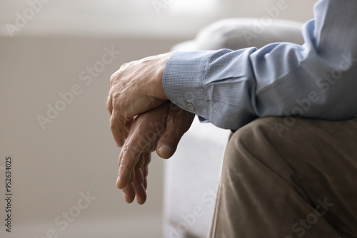 Cropped view, folded hands of unknown senior, man sitting on couch indoors. Close up shot of unrecognizable lonely retired male, deep in thoughts about senile diseases, loneliness in nursing house