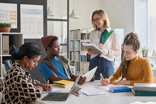 Young smiling female teacher enjoying class in school or college with students group sitting at table with books and computer