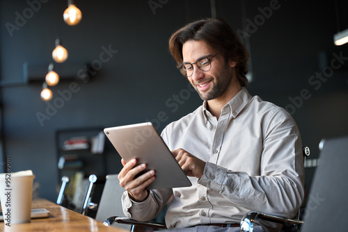 Happy young European business man entrepreneur holding tad computer at work. Male professional employee worker using digital tablet fintech device sitting in office checking financial online data.