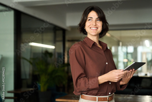 Happy young Hispanic professional business woman using tab standing in office. Happy businesswoman manager female executive leader holding tab at work looking away dreaming of future successful career