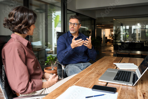 Business team of two busy professionals working together discussing corporate strategy. Male executive manager explaining corporate project plan to female partner colleague sitting at office desk.