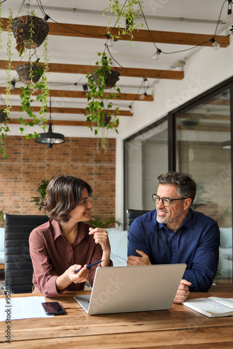 Two busy business team people using laptop computer working together sitting at office meeting. Happy young woman and mature man entrepreneurs executives smiling at work talking in workspace. Vertical