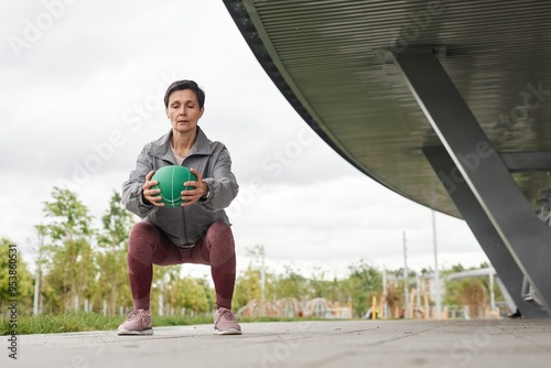 Full shot of fit senior woman holding ball in static squat position working out at city park against gray autumn sky, copy space