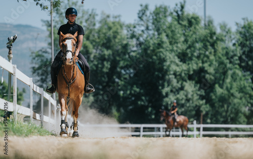 Man riding horse at equestrian facility on a sunny day. The scene features white fences and lush green trees in the background.