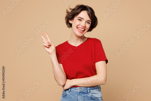 Young smiling cheerful fun cool happy woman she wearing red t-shirt casual clothes showing victory sign look camera isolated on plain pastel light beige background studio portrait. Lifestyle concept.