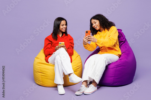 Full body two young smiling little kid teen girls women they wear red yellow hoodie casual clothes sit in bag chair hold use mobile cell phone isolated on plain purple background. Lifestyle concept.