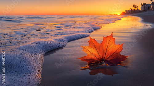 A colorful maple leaf lies on the beach, with waves lapping at its edge and the sun setting in the background