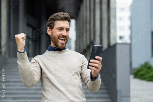 Man celebrating success while looking at smartphone. He is expressing happiness and excitement with fist pump. Urban background adds dynamic energy to his joyful reaction.