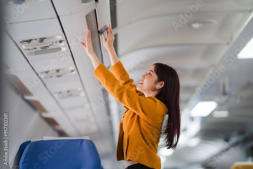 Ready for Takeoff: A young Asian woman stows her carry-on luggage in the overhead compartment of an airplane, her smile reflecting the excitement of upcoming travel. 