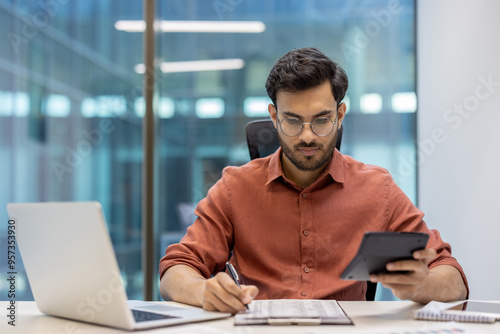 Serious concentrated hispanic businessman financier on paperwork inside office, man in business suit at workplace reviewing and reading papers, contracts and accounts reports