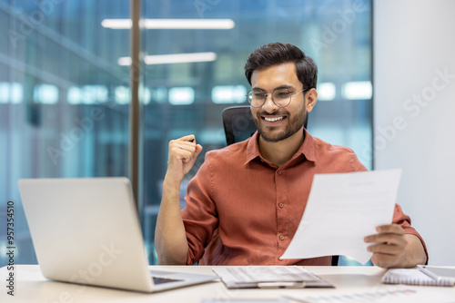 Confident businessman celebrates success at modern office desk with laptop and documents in hand, feeling accomplished during workday