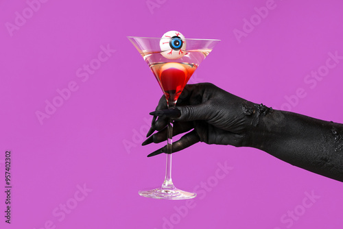 Black hand of witch with glass of cocktail and eye on purple background. Halloween celebration