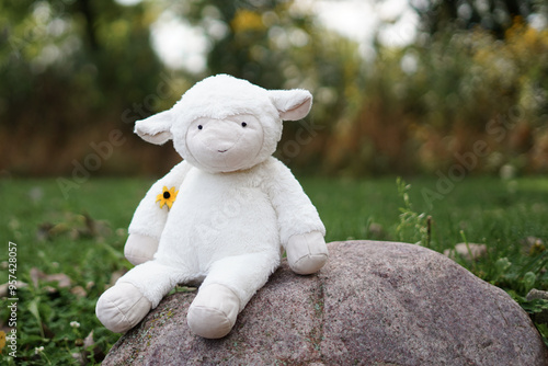 a cute plush lamb toy sitting on a rock in an outdoor setting, with a small yellow flower tucked under one arm