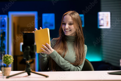 Upbeat gen Z content creator films video review of book using smartphone on tripod, presenting synopsis to audience. Media star shows new novel release to subscribers, recording with phone