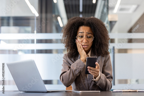 Shocked and scared young African American woman is in the office, sitting at the desk and looking at the mobile phone screen, holding her hand to her cheek