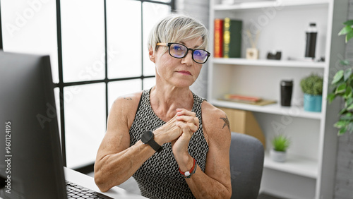 Mature woman with glasses and short grey hair contemplates in modern office setting.