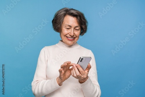 Happy aged caucasian woman grandmother holding smartphone using mobile online apps, browsing. Smiling old elderly lady texting sms message chatting with family on phone isolated over blue background.