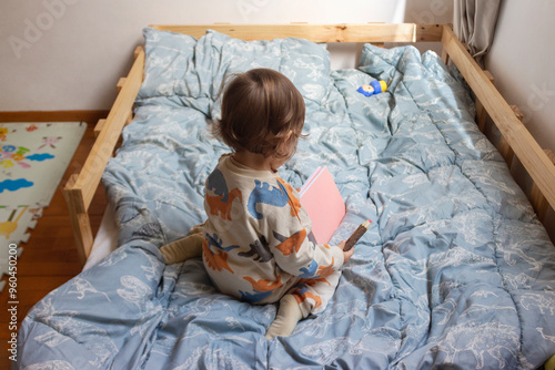 Bebé de un año sentado en su cama, él está pintando con sus colores sobre un cuaderno con hojas blancas, él está de espaldas, ángulo contrapicado, cubrecama azul