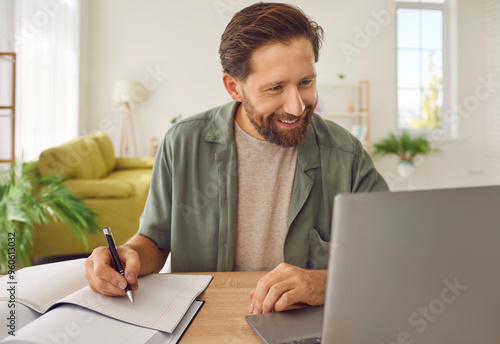 Happy adult man studying online. Smiling bearded male student in casual shirt sitting at desk at home, using laptop computer, watching lecture, learning new information, taking notes in paper notebook