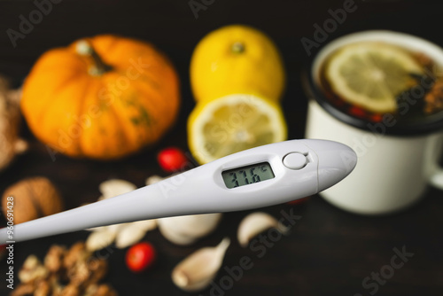 Cold and flu season, digital thermometer indicates high temperature 37.6, above wooden table with natural remedies, cup of herbal tea, lemon, garlic, pumpkin seeds, ginger and dog rose fruit