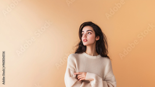 A pensive girl in a warm jumper against a beige wall points her finger sideways. Beautiful dark-haired girl in cosy jumper, copy space on the side