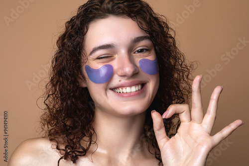 Smiling young girl with curly hair and purple under-eye patches, winking playfully while showing an OK sign. Skincare routine concept with vibrant energy and natural beauty on a beige background...