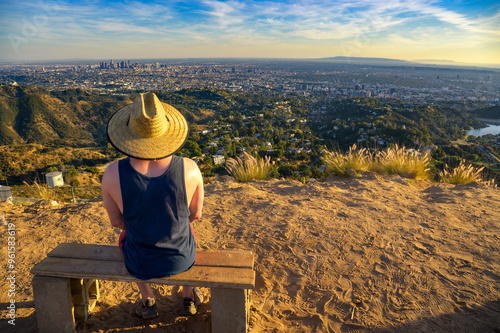 Tourist in a straw hat sits on a bench overlooking the Los Angeles skyline from Mount Lee at sunset.