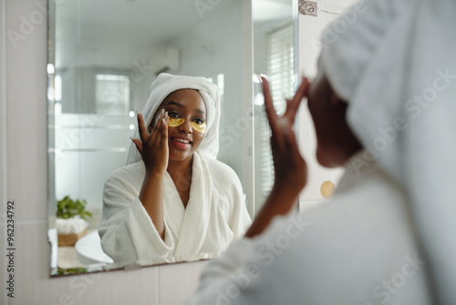 African American woman applying under-eye patches while looking in bathroom mirror, wearing bathrobe and towel on head. Reflecting in large, bright bathroom mirror while performing skincare routine