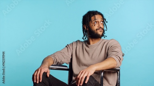 Bored Middle Eastern man with disability in wheelchair impatiently tapping fingers on knee, waiting. Annoyed person with paraplegia looking around, isolated over studio background, camera A