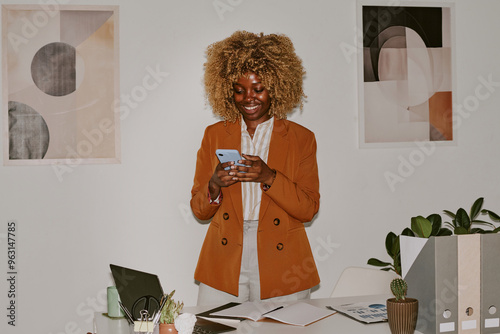 Blonde curly haired woman texting in social network and smiling widely while standing next to her worktable