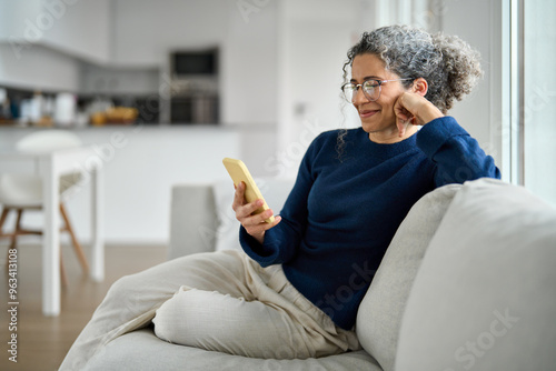 Happy middle aged older woman using smartphone sitting on couch at home. Smiling mature lady holding cellphone browsing internet, texting messages, doing online shopping on mobile cell phone on sofa.