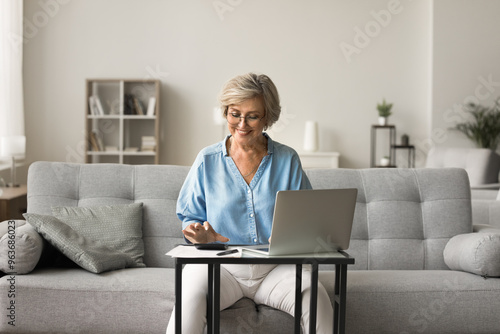 Mature woman use calculator while managing finances, sit on sofa at table with laptop and bills, counts incomes and expenses, smile, feel satisfied with money enough to make monthly household payment