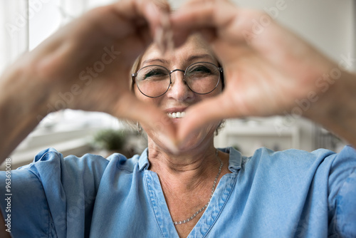 Mature woman wears eyeglasses looking at camera through joined fingers showing heart symbol, close up. Advertises eyewear store, laser vision correction, health check-up, cardio vitamins, sign of love