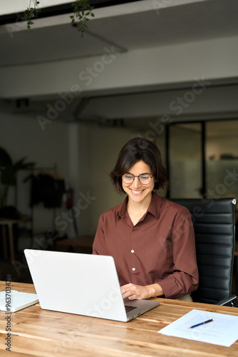 Busy young business woman executive using laptop in office. Smiling Hispanic businesswoman company worker sitting at work desk, professional female hr manager looking at pc computer at work. Vertical