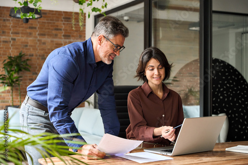 Two happy busy business leaders man and woman talking in office using laptop. Professional executive team people partners checking financial report working together on computer at corporate meeting.