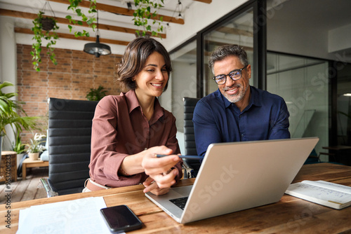 Business team of two executives working together using laptop in office. Happy young business woman manager showing project presentation to mature man client, investor or partner at work meeting.