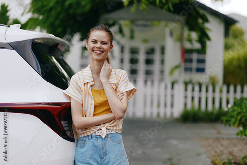 Smiling young woman leaning against a car in a sunny outdoor setting with a house in the background, wearing casual clothes and enjoying a bright day