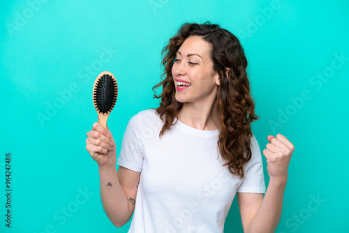 Young caucasian woman holding hairbrush isolated on blue background celebrating a victory