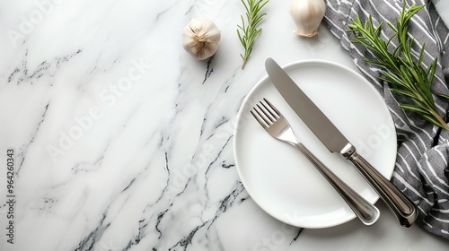 A white plate with a fork and knife on a marble surface, with rosemary sprigs and garlic cloves around.